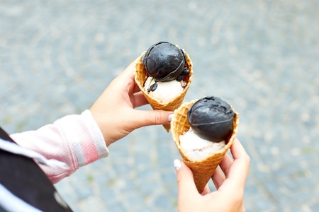 Female hand holding two charcoal and vanila ice cream in waffle cone on city background. Italian...