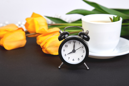 Black alarm clock near bouquet yellow tulips and cup of lemon tea on black background. Mother or Woman  Day. Greeting Card. Good Morning breakfast. Copy space. Spring.の写真素材