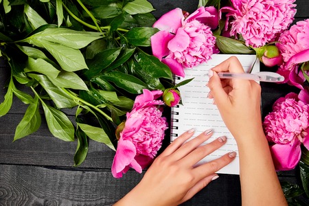 Woman hand writing diary, or letter, note. Peony flowers on black background. Planing to do list. Copy space.  Empty blank. Woman workplace. Good morning. Flat lay.の写真素材