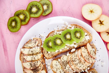 Healthy breakfast toasts with kiwi, apple, cottage cheese and chia seeds on white plate and pinkの写真素材