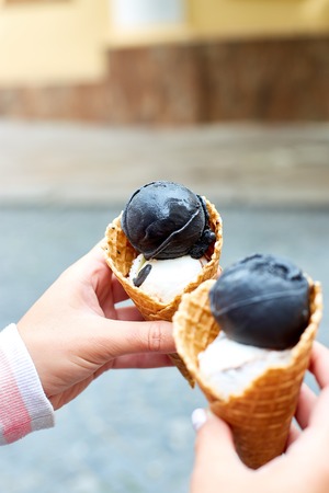 Female hand holding two charcoal and vanila ice cream in waffle cone on city background. Italian traditional ice cream gelato. Copy space.の写真素材