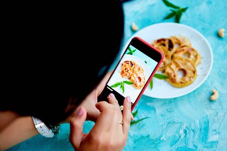 Hands with the phone close up pictures of food. Blogger girl. Food photo. Food blogger using smartphone taking photo. Grilled apples with cinamon on white plate on blue background.の写真素材
