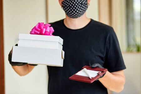 Courier man in black with protection medical mask and gloves contactless delivery presents, gift box...