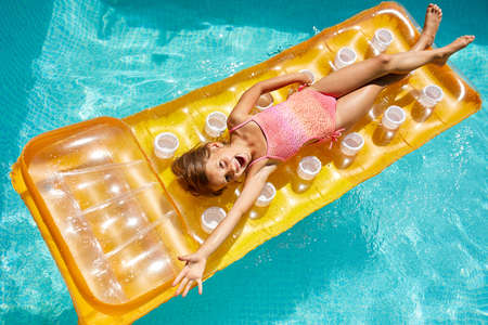 Happy little girl has fun in swimming pool, swims on inflatable yellow mattress on family vacation,...