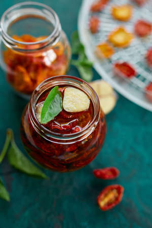Homemade sun dried tomatoes with herbs, garlic in olive oil in a glass jar on dark green background....