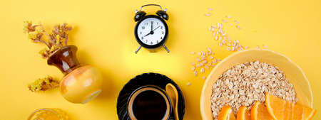 Banner of Morning coffee, granola breakfast with fruit near black alarm clock, vase flower on yellow background. Flat lay. Copy space. Top view.の写真素材