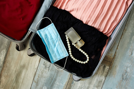 Packing suitcase at home with woman items, accessories and protection mask during pandemic in...