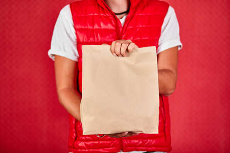 Courier, delivery man in red vest uniform delivers online purchases in brown paper bags to the door, isolated on red background studio.の写真素材
