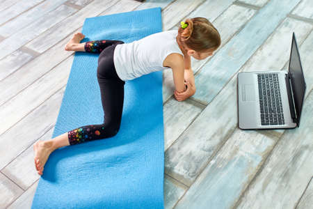 Little girl practicing gymnastics at home, watching online video on laptop. Social distance, online...
