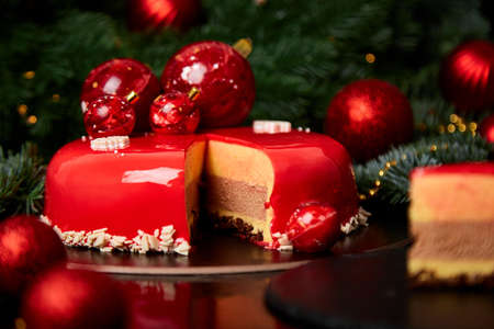 Mousse Christmas pastry cake dessert covered with red mirror glaze with new year decorations on garland lamps bokeh dark background, Modern european cake Christmas theme.の写真素材