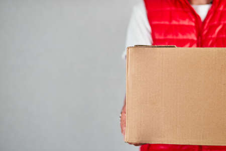 Delivery man employee in red vest uniform hold empty cardboard box isolated on grey background studio, Service quarantine pandemic, Contactless delivery.の写真素材