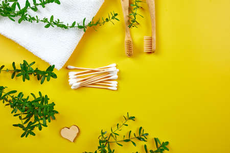 Bamboo toothbrushes and ear sticks with towel, and green leaves on yellow paper background,...