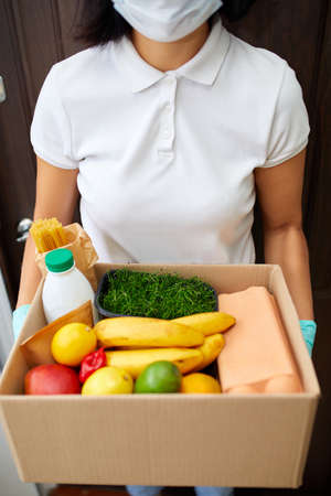 Courier with packing box with food, contactless delivery, Service quarantine pandemic, Volunteer woman in white protective mask and gloves delivery donation box at home.の写真素材