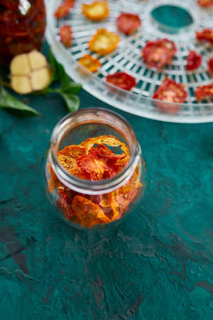 Homemade sun dried tomatoes with herbs, garlic in olive oil in a glass jar on dark green background....