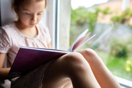 Cute little girl reading book at home, at windowsill, Child studying at home, quarantine, stay home, life style.の写真素材