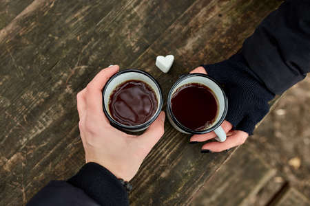 Two cups of tea in nature in the hands of couple in black gloves on wooden background, love, heart of marshmallow, travel concept, Lifestyle moment at nature, Valentine day, Copy space, top view.の写真素材