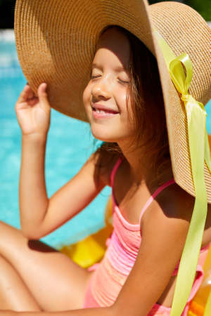 Portrait of little girl in hat relaxing in swimming pool, swims on inflatable yellow mattress and...