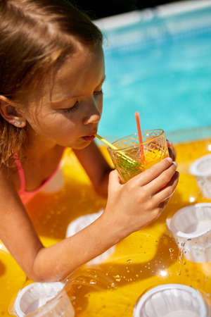 Little girl relaxing in swimming pool, enjoying suntans, drink a juice on inflatable yellow mattress...
