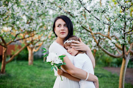 Cute daughter and mother hugging in blossom spring garden Happy woman and child, Wearing white dress outdoors, Spring season is coming. Mothers day holiday conceptの写真素材