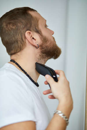 Handsome man in white t-shirts cutting beard, moustache personally himself with trimmer at home. Self Care during quarantine isolation. Stay home.の写真素材
