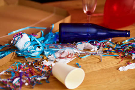 Messy wooden table with pizza box, empty blue bottles, wine glass and party decorations at morning after party celebration, After party chaos, Celebration leftovers.の写真素材