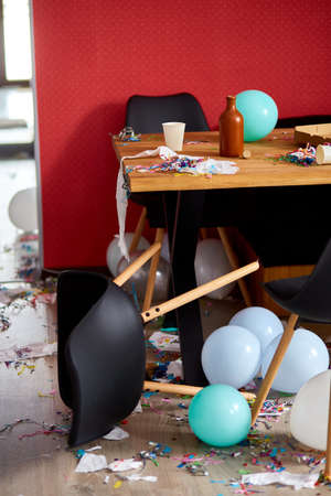 After party chaos, messy in livving room at home, table with pizza and champagne glasses covered with confetti and ballons, chair on the floor at morning after party celebration.の写真素材
