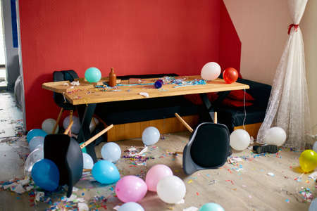 After party chaos, messy in livving room at home, table with pizza and champagne glasses covered with confetti and ballons, chair on the floor at morning after party celebration.の写真素材