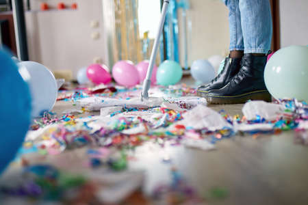 Woman with pushbroom cleaning mess of floor in room after party confetti, morning after party celebration, housework, cleaning serviceの写真素材