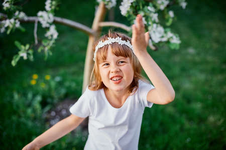 Portrait  of funny kid girl 5-6 year old holding flower standing in blossom spring garden, Wearing...