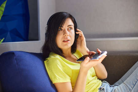 Cheerful woman in casual clothes with headphones and smartphone, record to podcast on sofa at home....