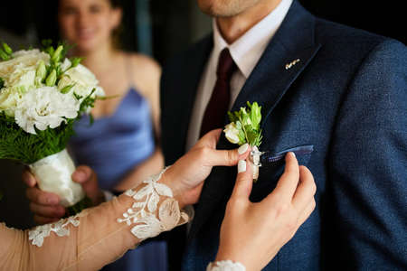The bride's hand puts on a boutonniere flower on the groom's jacket. Bride puts a buttonhole on a grooms suitの写真素材