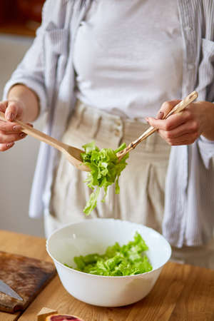 Woman is preparing vegetable salad in the kitchen, mixing leaf of salad and vegetables ina white bowl, Healthy food concept, vegan or diet.の写真素材