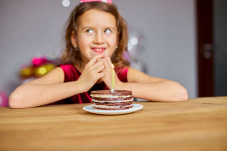 A little girl is wearing a birthday hat makes a wish , looking at a Birthday Cake, with glowing...