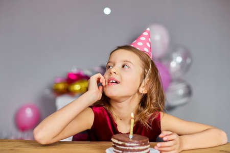 A little girl is wearing a birthday hat makes a wish , looking at a Birthday Cake, with glowing...