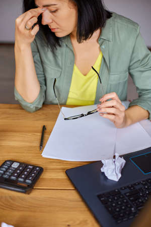 Stylish brunette business woman takes off glasses working in modern workplace, overworked female feeling eye strain after using computer working home. Headache, visual fatigue and eye strain concept.の写真素材