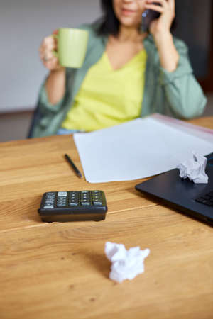 Stylish brunette woman in glasses sitting at wooden table with notepad and having phone call, drink...