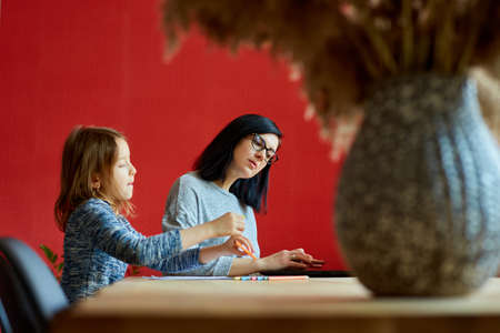 Mother working in her home office on a laptop, her daughter sits next to her and draw, Woman freelance, remote work and raising a child at workplace.の写真素材