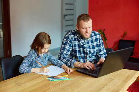 Father working in his home office on a laptop, her daughter sits next to her and draw, Man...