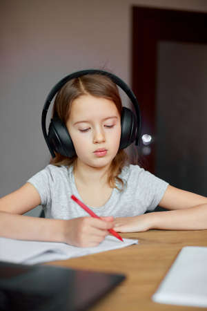 Cute little girl with headphones using laptop to study at home, writing, answer, online learning, education, social distance, quarantine.の写真素材