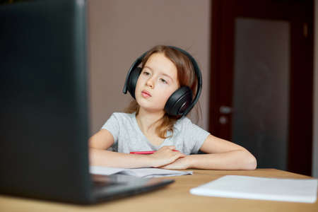Cute little girl with headphones using laptop to study at home, writing, answer, online learning, education, social distance, quarantine.の写真素材