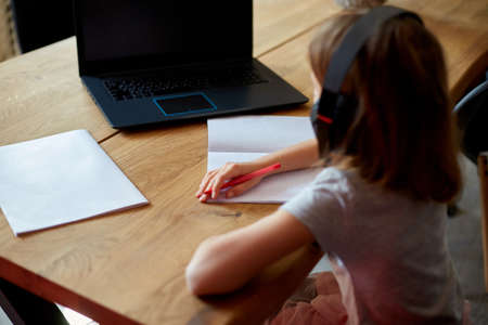 Cute little girl with headphones using laptop to study at home, writing, answer, online learning, education, social distance, quarantine.の写真素材