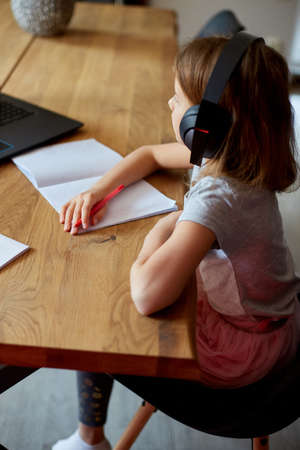 Cute little girl with headphones using laptop to study at home, writing, answer, online learning, education, social distance, quarantine.の写真素材