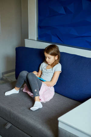 Cute little girl sitting on couch, Kid addicted to technology, enjoying playing online game on digital tablet computer, using applications, web surfing information at home,の写真素材