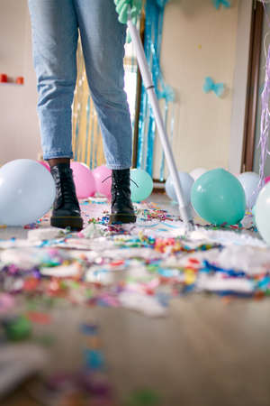 Woman with pushbroom cleaning mess of floor in room after party confetti, morning after party...