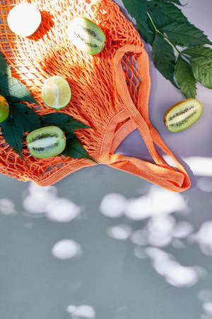 Flat lay of eco friendly mesh shopping bag With Fruits lime and kiwi on grey background in sunlight,...