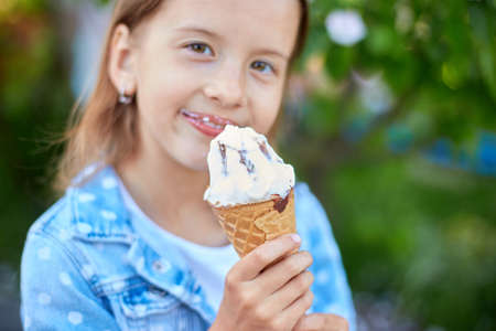 Cute girl with italian ice cream cone smiling and looking at camera while resting in park on summer day, child enjoying ice cream outdoor, happy holidays, summertimeの写真素材