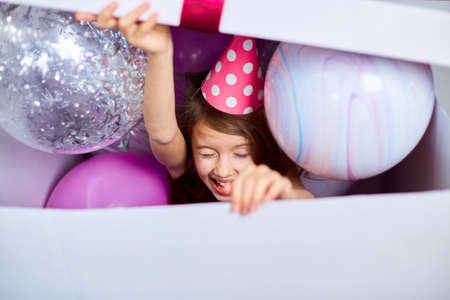 Cute, joyful little girl in pink dress looks out of the big present gift box with balloons at home birthday party streamers, Happy birthday. Celebratingの写真素材
