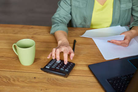 Close up of woman sit calculate expenses on calculator at wooden table, modern workplace, freelancer working at home. Female busy manage family budget account expenditures.の写真素材