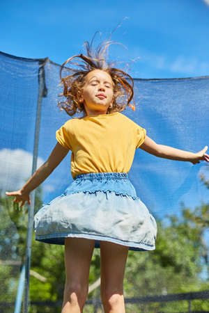 A young girl jumping up and down on her trampoline outdoors, in the backyard of the house on a sunny summer day, summertime vacation.の写真素材