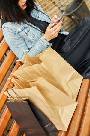 Woman sitting on the bench in the park withpaper shopping bags after shopping, holding in hands...